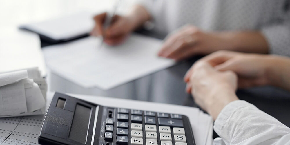 Woman accountant using a pen and laptop computer while counting and discussing taxes with a client, focus on the calculator. Business audit and finance concepts.
