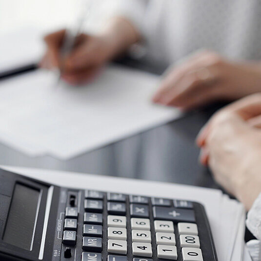 Woman accountant using a pen and laptop computer while counting and discussing taxes with a client, focus on the calculator. Business audit and finance concepts.