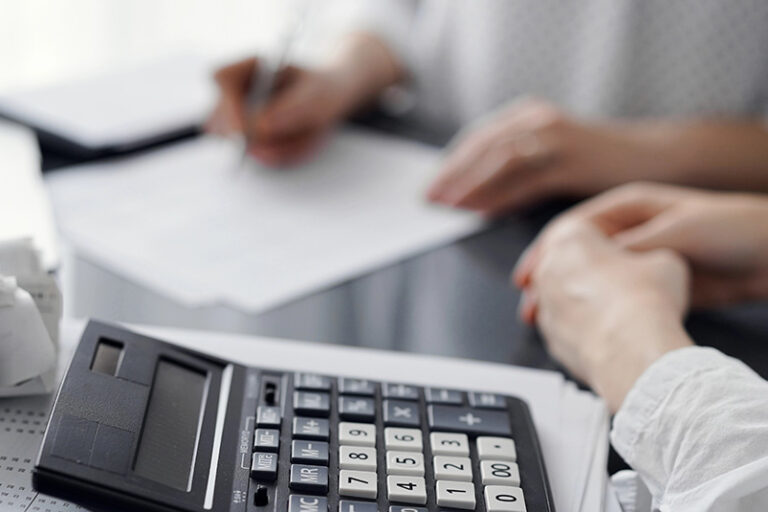 Woman accountant using a pen and laptop computer while counting and discussing taxes with a client, focus on the calculator. Business audit and finance concepts.